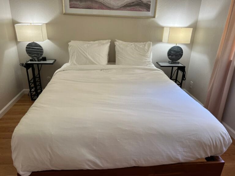 A neatly made bed with white bedding, two pillows, and two bedside tables with matching lamps in a simply decorated bedroom—perfectly prepared after a Deep Cleaning service in San Jose.