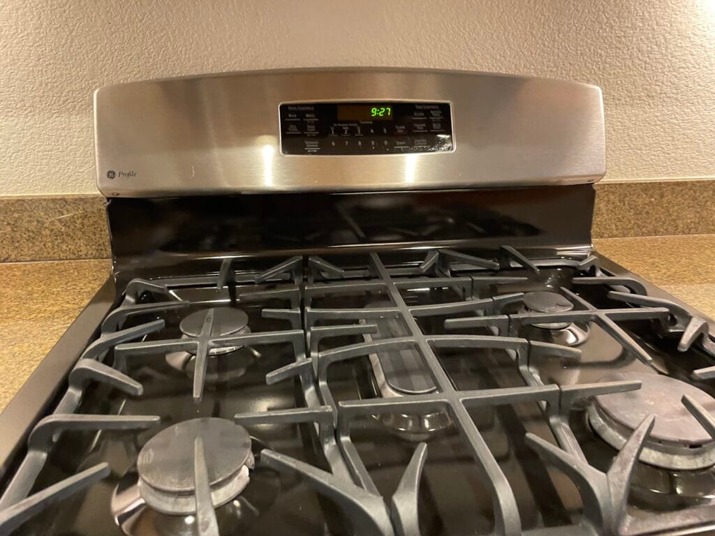 Stainless steel gas stove with four burners and an electronic control panel displaying 5:27, set against a beige wall and brown countertop. Ideal for easy regular cleaning, perfect for busy San Jose kitchens.