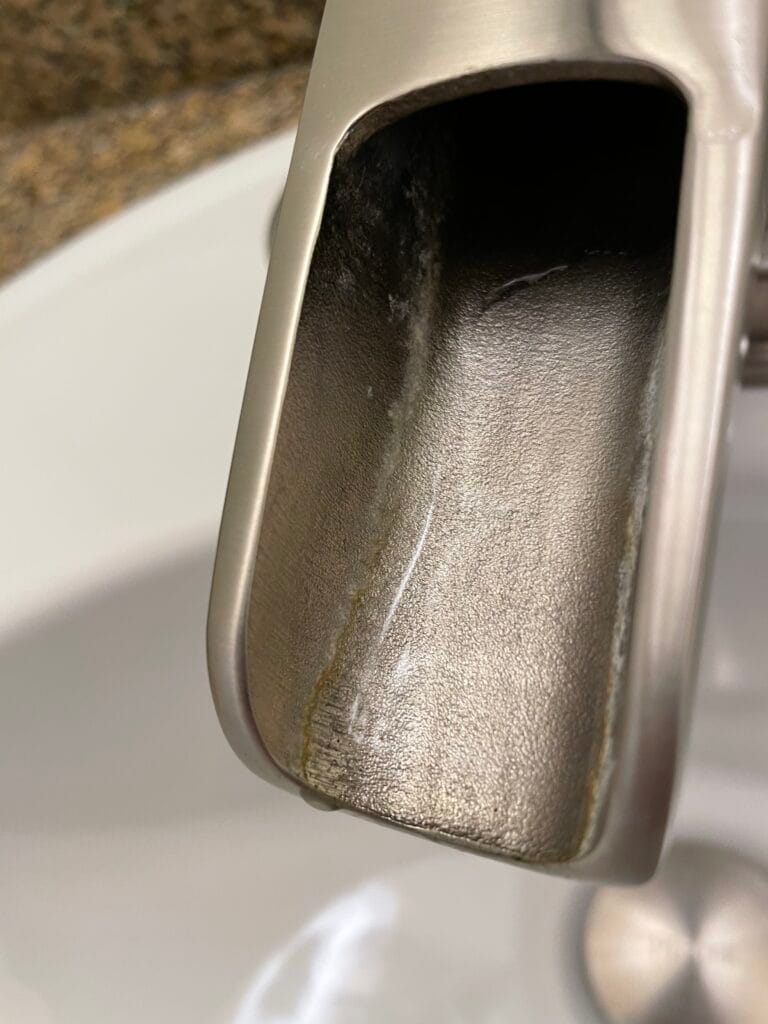 Close-up of a metal faucet spout with visible mineral buildup and water stains above a white sink, highlighting the need for regular cleaning to maintain its shine.